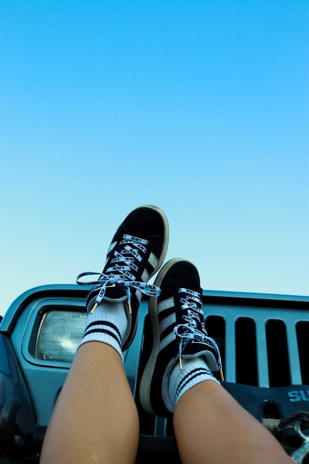 Feet up on a jeep against blue sky, black sneakers laced with white LACIE All-Over Laces